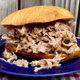 Pork sandwich on a blue plate atop a red checkered tablecloth, with a wooden fence in the background